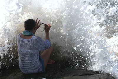 Rear view of woman sitting while waves splashing on rock