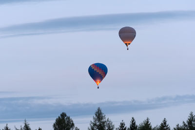 Two hot air balloons float in the cloudy sky. in the foreground, treetops are visible 