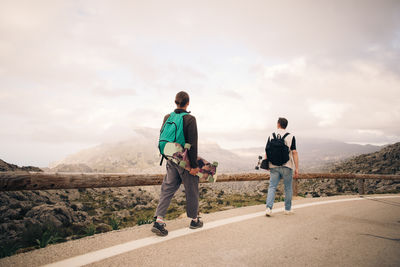Young male friends looking at mountains while walking on road holding skateboard during vacation