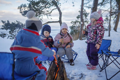 Family winter picnic. happy friends sit around campfire on the shore of frozen lake in forest