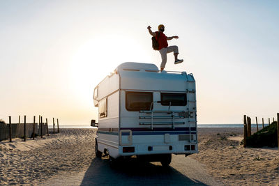 Cheerful male explorer standing on roof of camper parked on seashore and enjoying with outstretched arms and raised leg against sundown sky