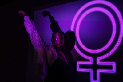 Trendy black model raising arm and looking away while standing near neon female symbol glowing with purple light