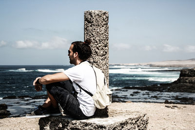 Side view of mid adult man with backpack sitting on rock at beach against sky during sunny day