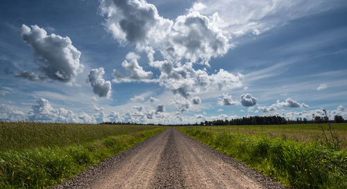 Dirt road amidst green field against cloudy sky during sunny day