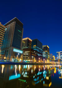 Illuminated buildings by river against blue sky at night