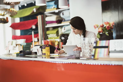 Female owner using digital tablet while writing on note pad at checkout counter in store