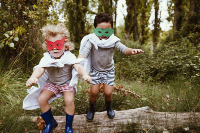 Playful boys wearing masks and capes jumping together in forest