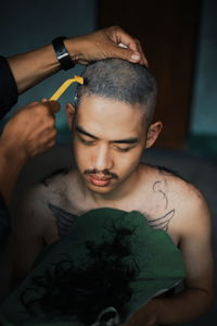 Cropped hands of person shaving shirtless man head at home