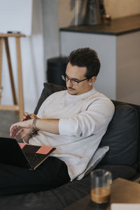 Young businessman checking time on wristwatch while sitting with laptop at office