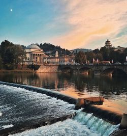 Buildings by lake against sky during sunset