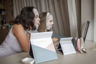 Mother and daughter applying face mask by mirror and laptop at home
