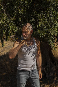 Adult man in white tank top and jeans standing with olive tree in summer