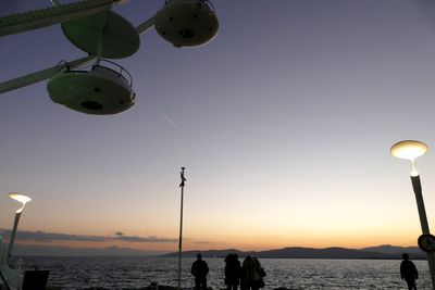 People at sea shore by ferris wheel against sky during sunset
