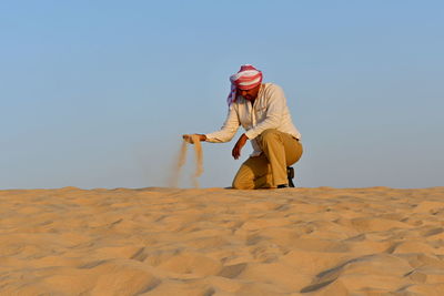 Full length of man holding sand while kneeling on desert against sky