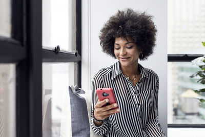 Happy businesswoman using smart phone against window in office