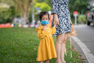 Little girl holding mother's hand wearing healthy face mask stand along the road.