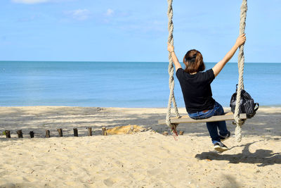 Boy sitting on swing at beach against sky