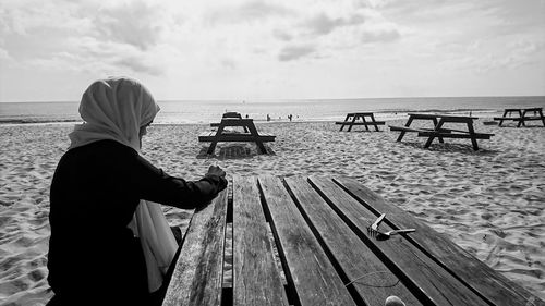 Woman sitting on picnic table at beach against sky