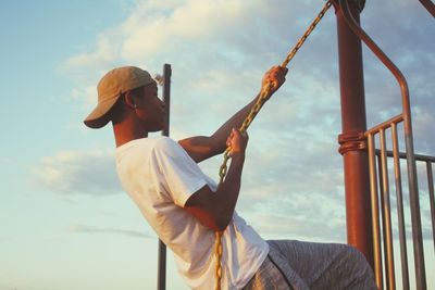 Man hanging from chain against sky