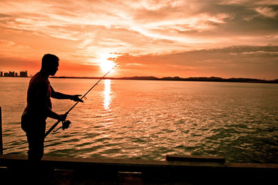 Silhouette man fishing in sea against sky during sunset