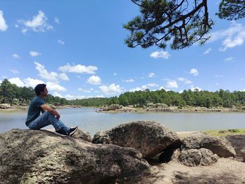 Man sitting on rock by lake against sky