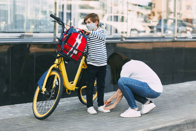 A mother adjusts her son's sneakers before riding a bike to school. a boy wearing a mask