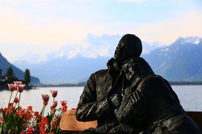 Human statues on bench at lakeshore against mountains