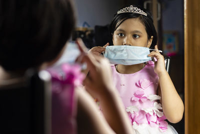 A cute indian girl child in red dress holding surgical nose mask in front of mirror