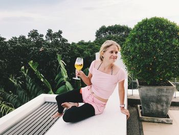 Woman having wine while sitting on wall against sky