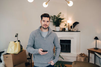 Portrait of smiling man holding mobile phone while standing with hand in pocket at home
