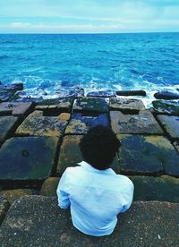 Rear view of man sitting on retaining wall sitting by sea against sky