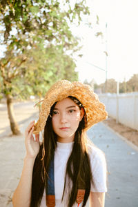 Portrait of young woman in hat standing on sidewalk against trees