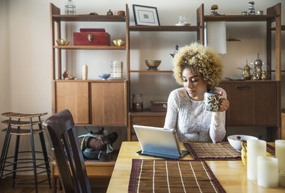 Smiling woman using laptop while holding coffee cup at table in house