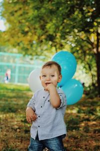 Portrait of cute baby boy with balloons standing against trees