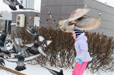 Side view of girl playing with pigeons during snowfall in city