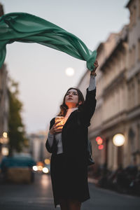 Woman moving green scarf while holding illuminated jar in city at dusk