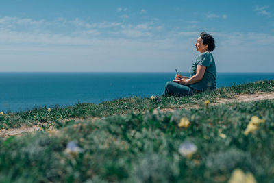 Adult woman thoughtfully writes plans diary on a sunny day on a mountain against backdrop of the sea