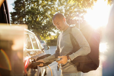 Man charging car while using mobile phone at electric station on sunny day