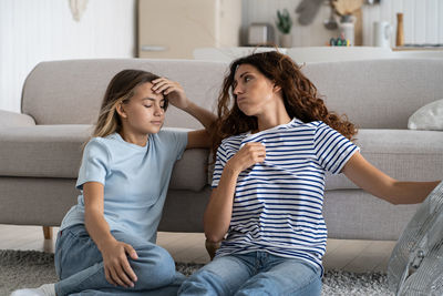 Sweaty tired woman sits on floor next to sofa and fan looking back at teenage girl after summer walk