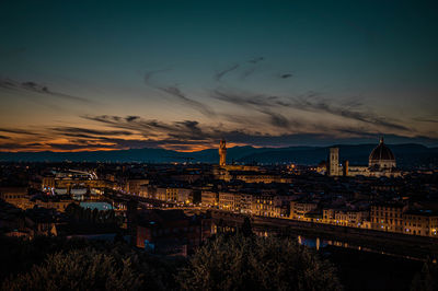 Illuminated bridge over river by buildings against sky at sunset
