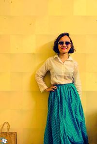 Young woman wearing sunglasses standing against yellow wall in an indo western attire