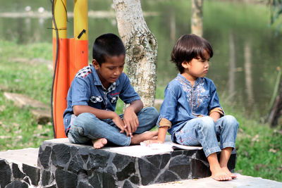 Boys reading book while sitting on seat against lake