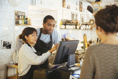 Young employee standing by female cashier using computer with customer at checkout counter in delicatessen