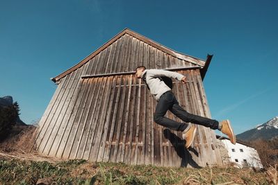 Low angle view of man levitating by barn against blue sky