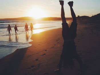 Full length of silhouette man doing handstand at beach against bright sky during sunset