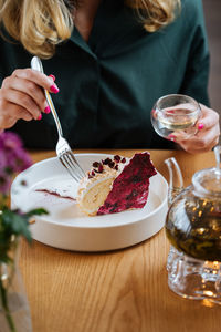Crop female in dark green blouse sitting at wooden table eating delicious cake with red berries served in white ceramic plate and drinking herbal tea