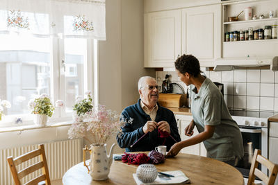 Female caretaker talking with senior man knitting yarn while sitting at table in kitchen