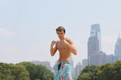 Portrait of young shirtless man standing against sky in city