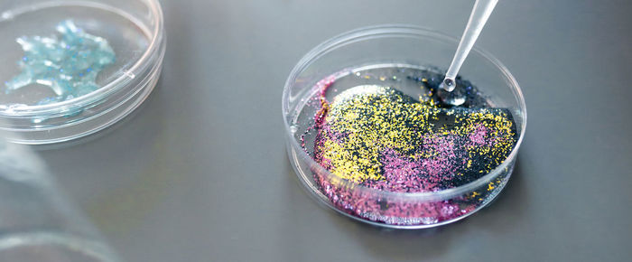 Drop of liquid falling from from pipette over petri dish with mix of colorful glitters in laboratory