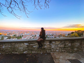 Rear view of woman sitting on retaining wall against sky during sunset